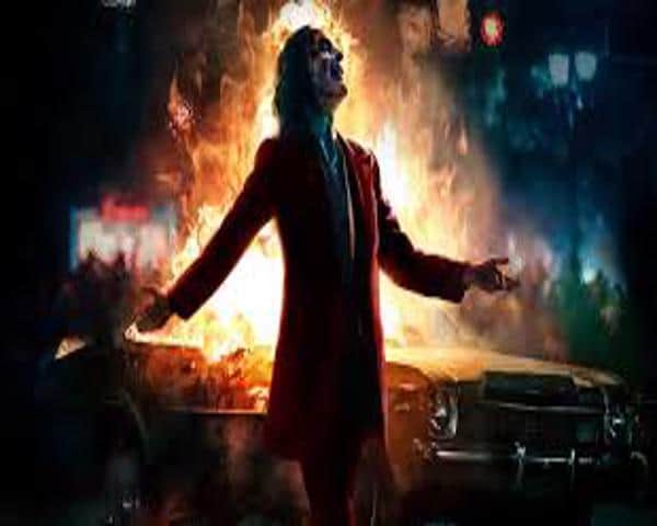 Joaquin Phoenix as the Joker, arms spread, in front of a burning car
