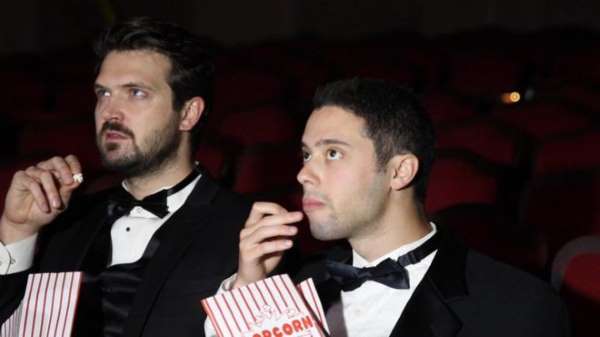 Two men in tuxedos eat popcorn in an empty movie theater