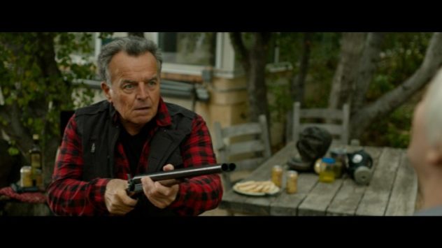 Still from Beast Mode: an older man in a plaid shirt aims a shotgun in a backyard