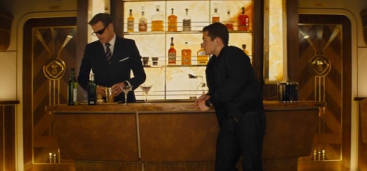 Still from Kingsman: a man in glasses mixes a martini at a bar as a younger man leans on the counter