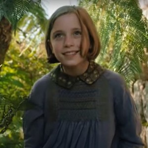 Still from The Secret Garden (2020): a smiling girl in a gray dress among lush ferns