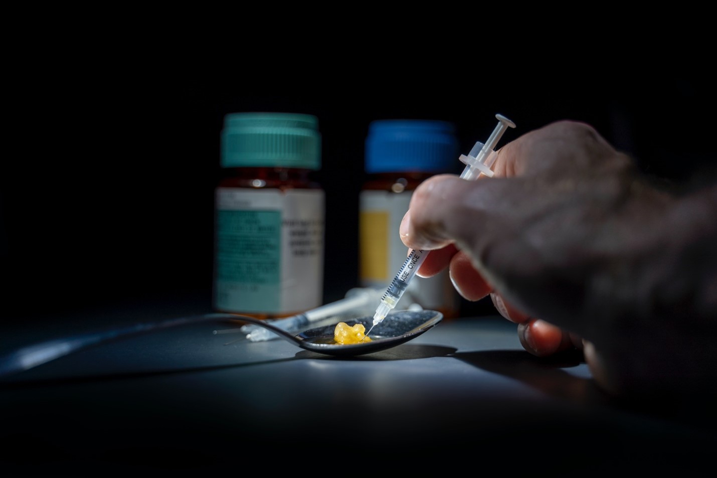 A hand holding a syringe over a spoon beside pill bottles, in the dark