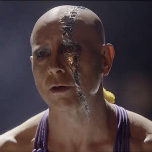 Still from I'm An Electric Lampshade: a bald performer with cracked, metallic face makeup on stage