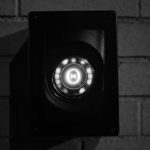 Still from Tes Yeux Mourants: a glowing round light in a black panel on a brick wall
