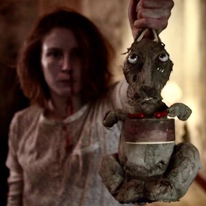 Still from Caveat: a bloodied woman beside a creepy stuffed rabbit toy held up by a hand