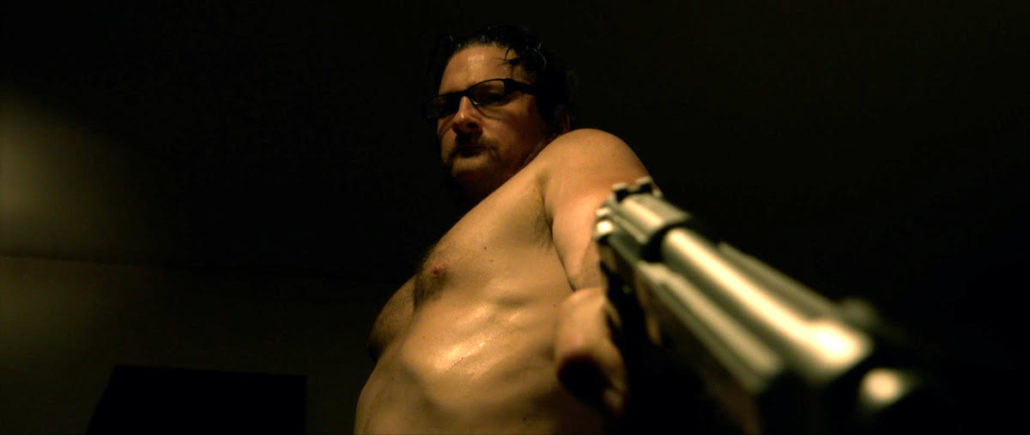 Still from Dreaming Hollywood: a shirtless man in glasses points a pistol at the camera