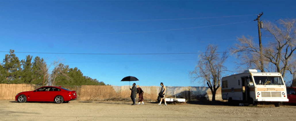 Still from Dreaming Hollywood: figures with an umbrella walk across a dusty lot by a red car and an RV