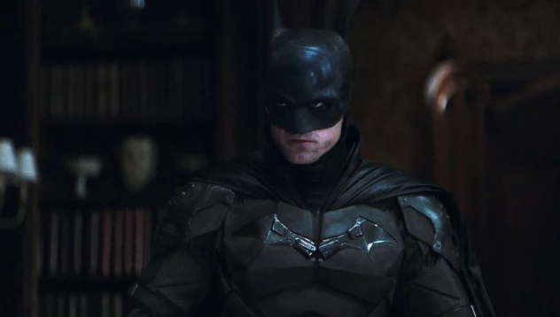Still from The Batman: Batman in his dark armored suit stands in a shadowy, book-lined room