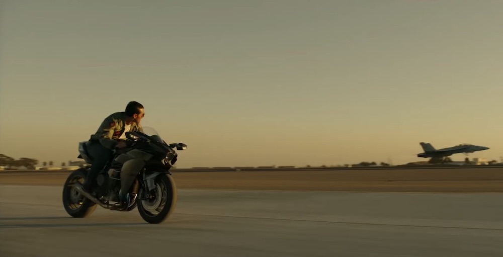 Still from Top Gun: Maverick: Maverick rides a motorcycle alongside a fighter jet on the runway at dusk