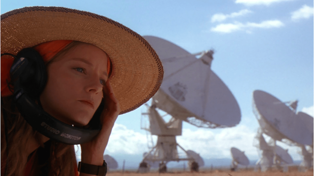 Still from Contact: a woman in a straw hat and headphones listens beside a field of radio telescope dishes
