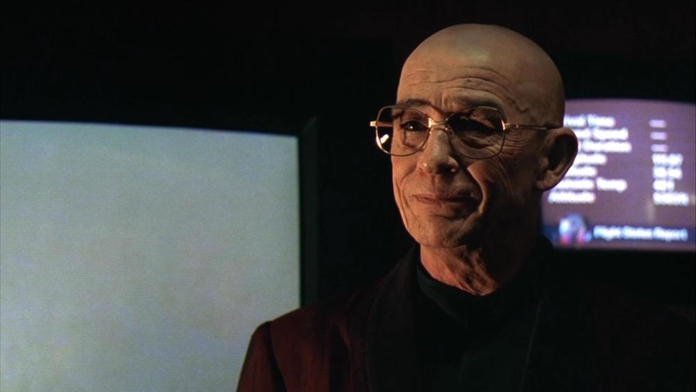 John Hurt as S.R. Hadden in Contact, bald and wearing glasses in a dim room