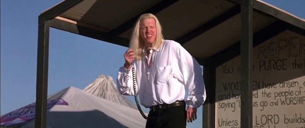 Still from Contact: Jake Busey as a long-haired preacher on a phone at a protest
