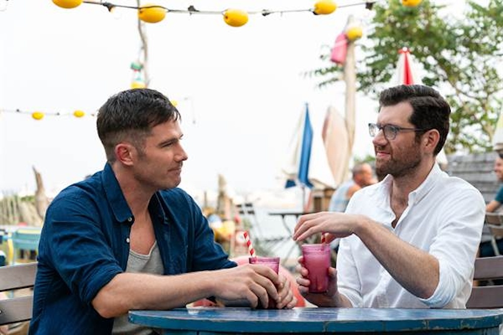 Still from Bros: two men chatting over drinks at an outdoor café table
