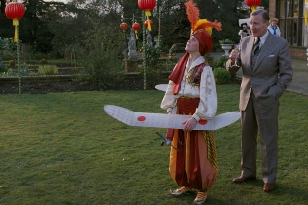 Empire of the Sun (1987): a boy in a feathered costume holds a model plane as a man in a suit watches