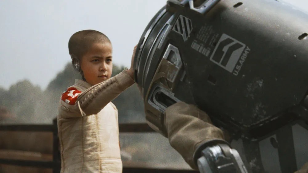 Still from The Creator: a young robot child with a shaved head touches a military robot's head