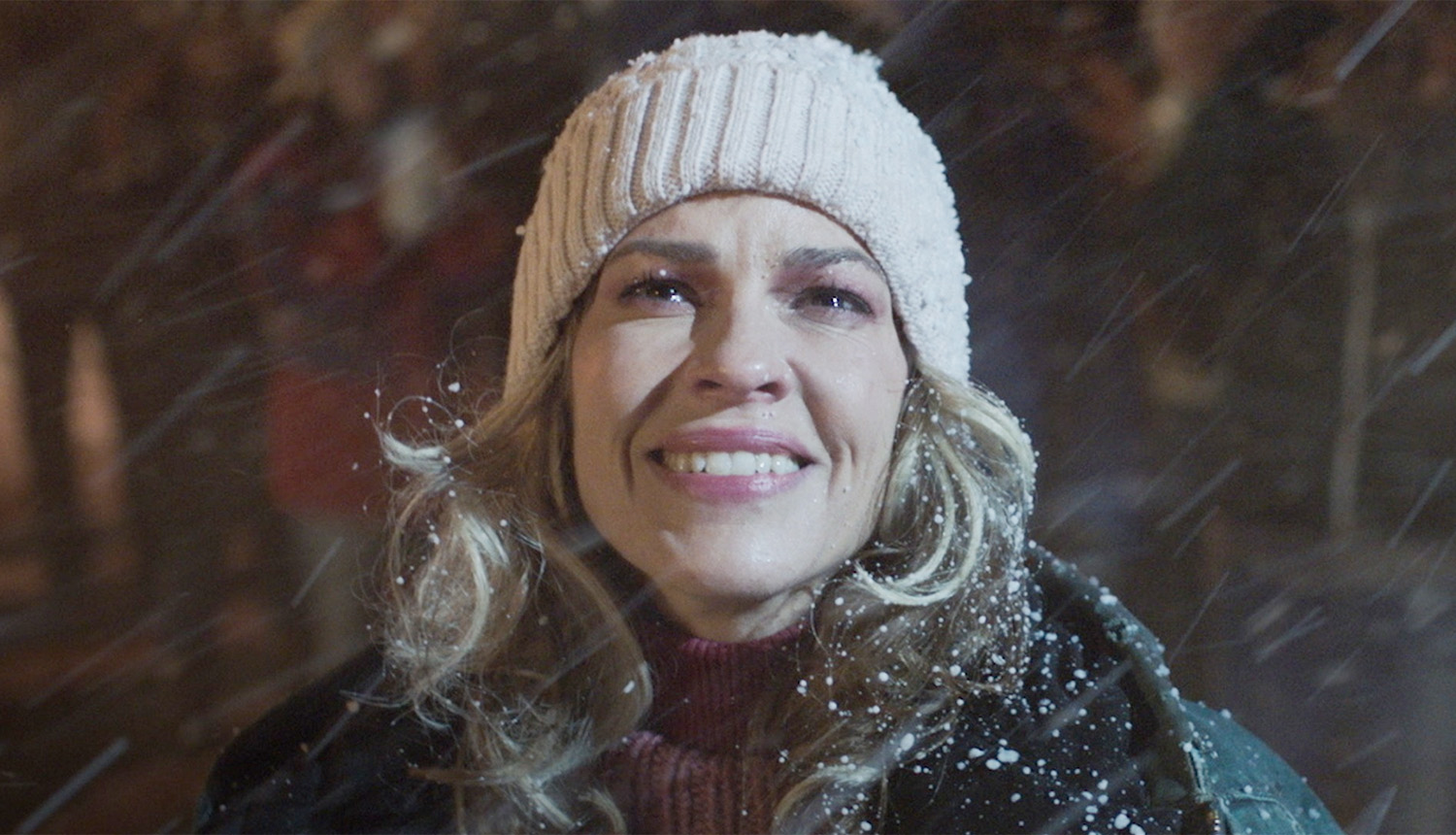 Still from Ordinary Angels: a woman in a white knit hat smiles up into falling snow