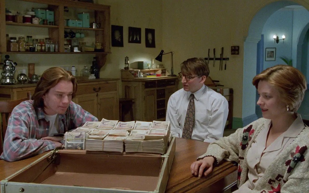 Still from Shallow Grave: three flatmates sit at a kitchen table around a case full of cash