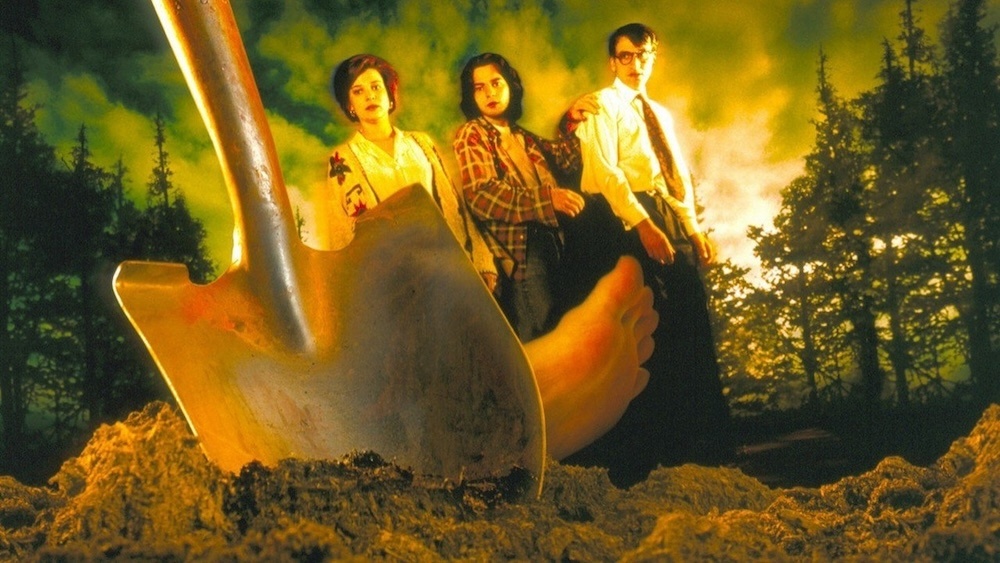 Shallow Grave promo still: three flatmates stand behind a shovel and a bare foot sticking out of fresh earth