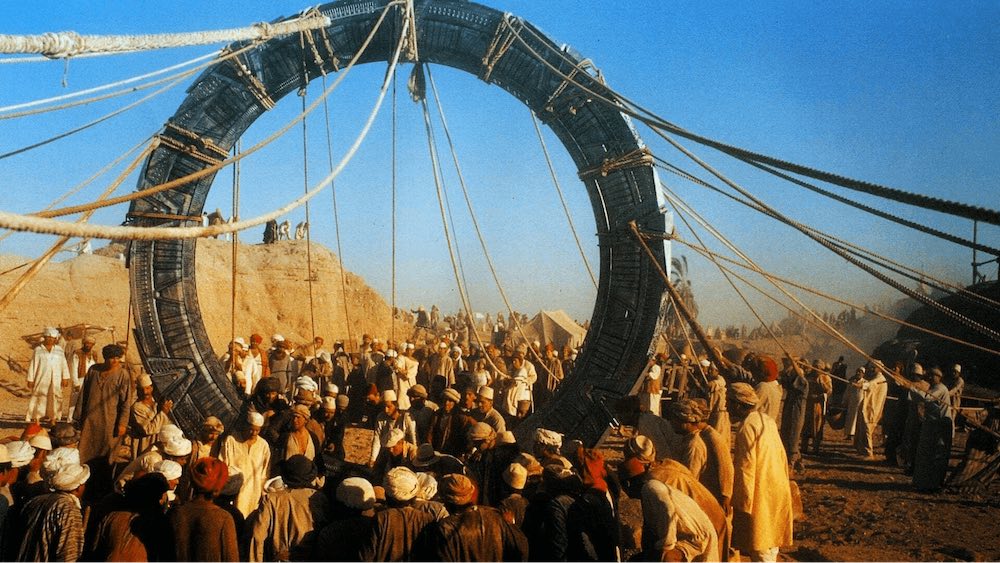 Still from Stargate: a crowd in desert robes hauls on ropes to raise the giant stone Stargate ring