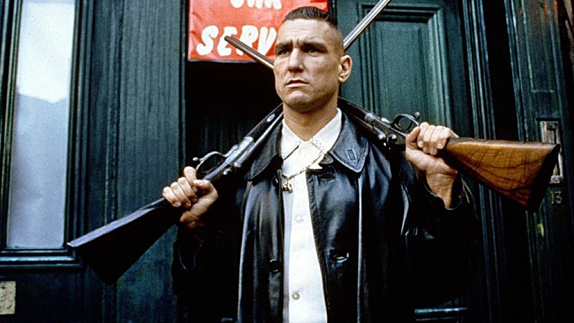 Vinnie Jones in a leather coat holding a shotgun over each shoulder