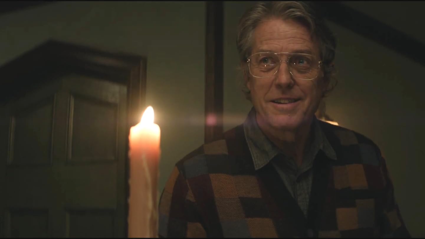 Hugh Grant in glasses and a patterned cardigan beside a candle in Heretic
