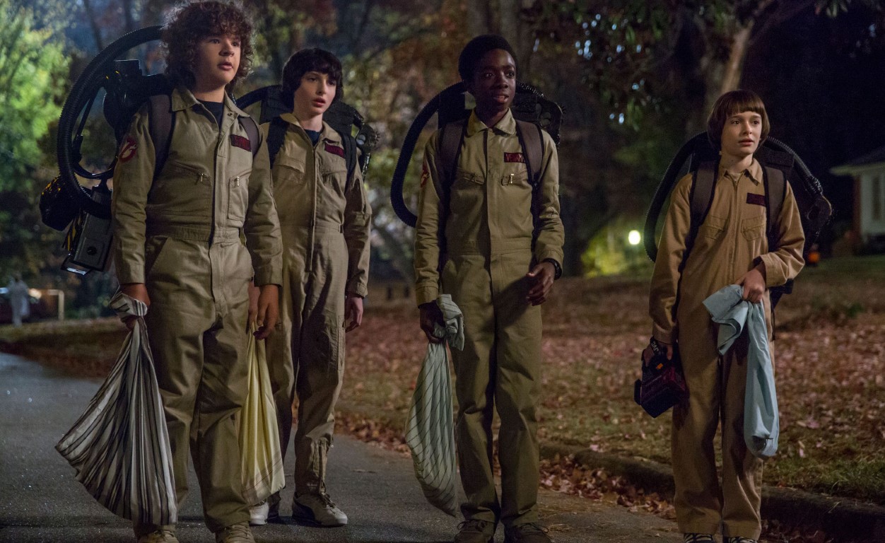 Still from Stranger Things season 2: four boys in Ghostbusters jumpsuits with proton packs trick-or-treat