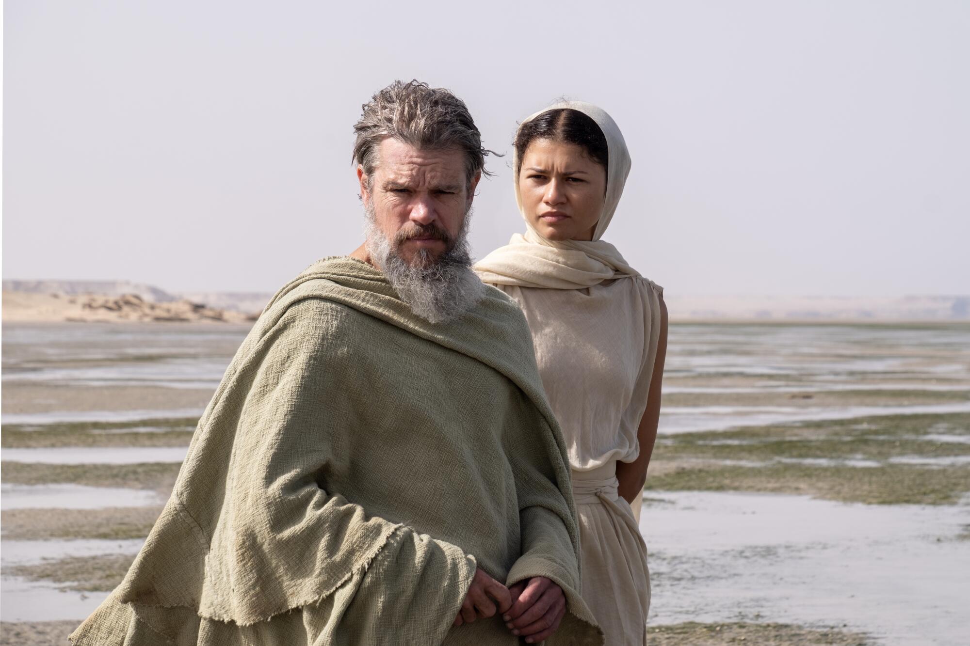 Still from The Odyssey: a gray-bearded man in a rough cloak and a young woman in a headscarf on tidal flats