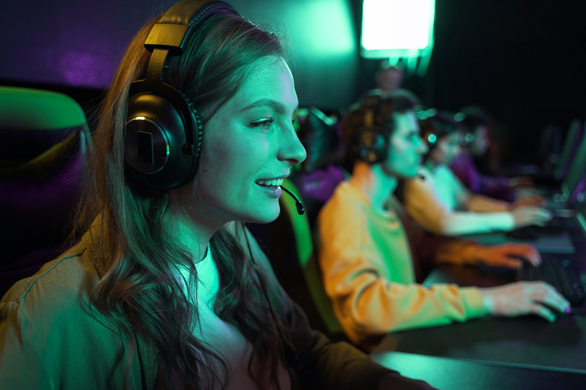 A smiling young woman in a headset plays games in a neon-lit room full of other gamers