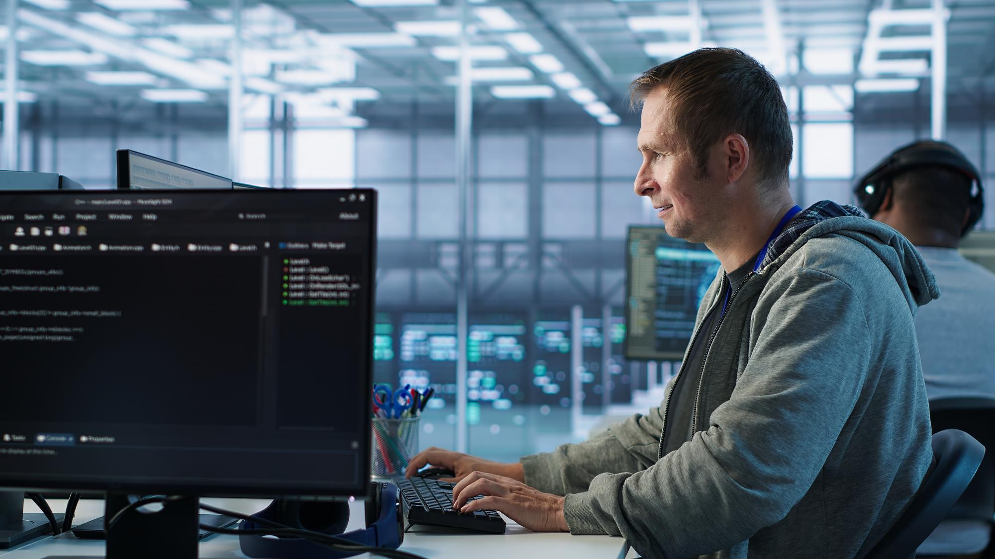 A man in a hoodie works at a computer showing code in a large open office lined with server screens